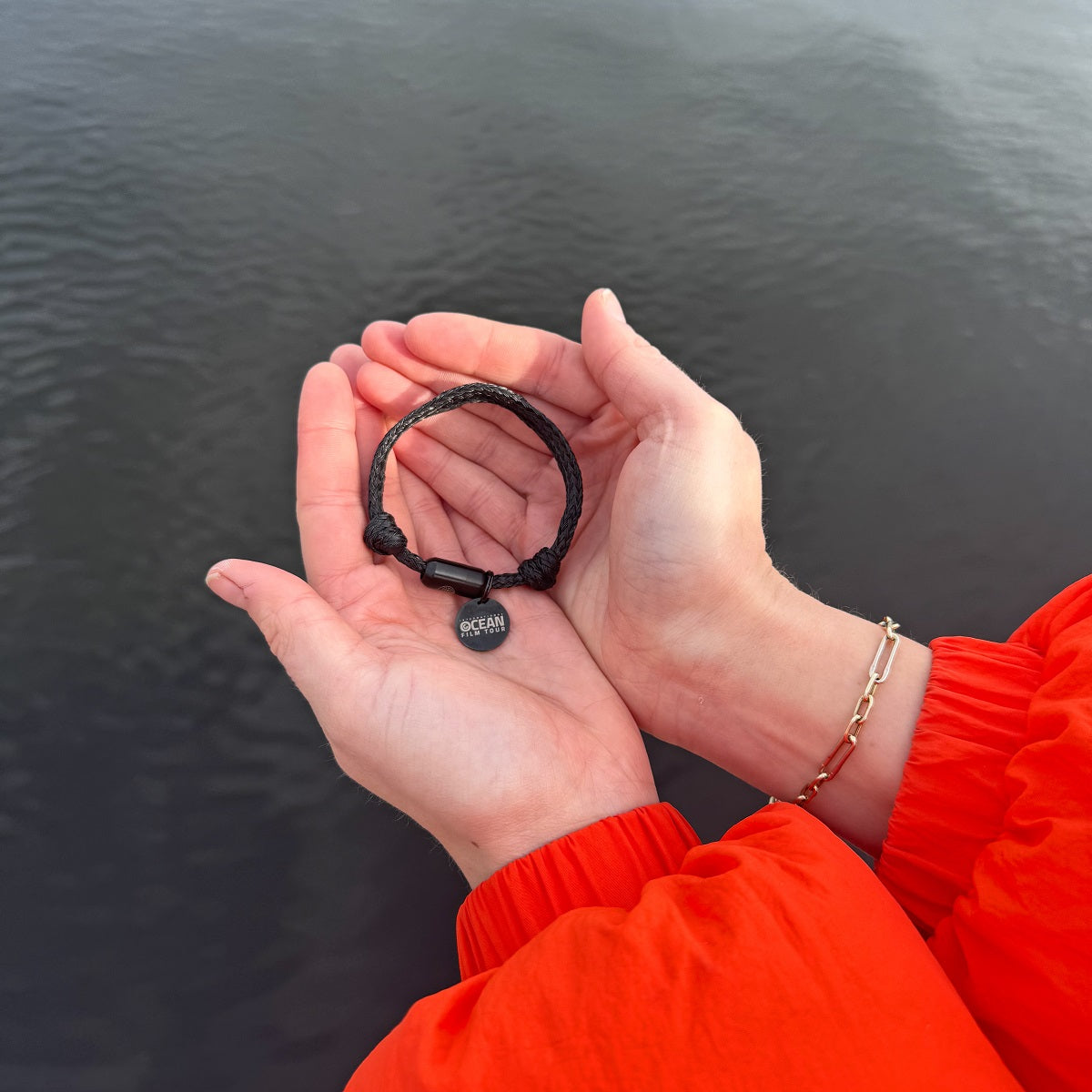 Person hält das Int. Ocean Film Tour 2025 Upcycling Armband in schwarz in die Kamera mit einem schwarzen Edelstahlanhänger und der Gravur Ocean Film Tour, im Hintergrund die Alster