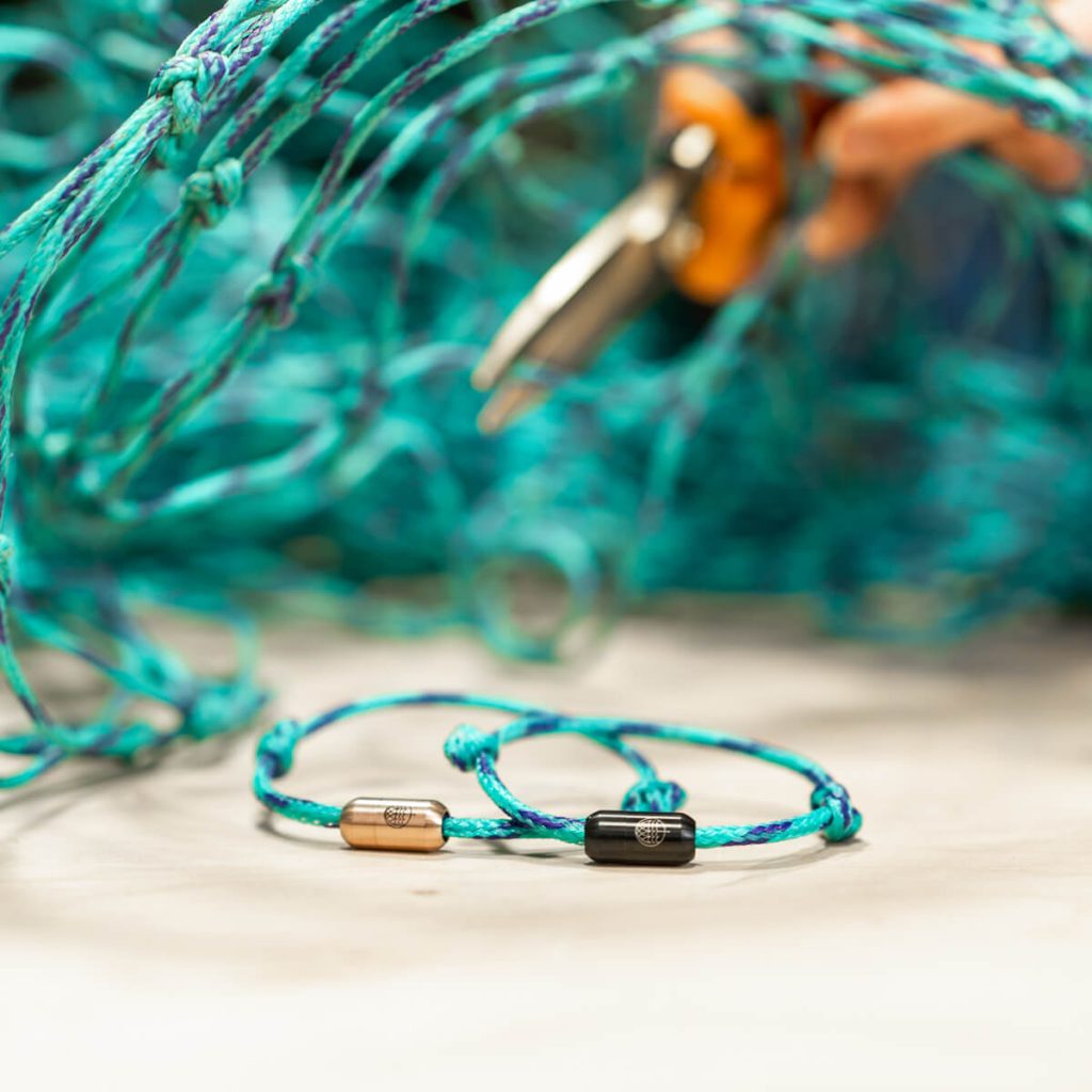Zwei fertige Bracenets liegen bei der Fertigung in Handarbeit auf dem Tisch, altes Fischernetz wird im Hintergrund zugeschnitten.