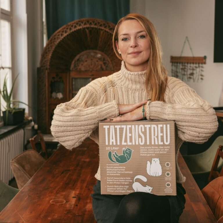 Madeleine sitzt in der Küche von Bracenet mit einem Karton TATZENSTREU auf dem Schoß und lächelt in die Kamera