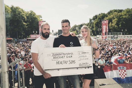 BRACENET Gründer:innen stehen vor einem großen Publikum mit einem Mitglied von Healthy Seas und halten gemeinsam einen großen Spendencheck in der Hand mit einer Spende über 20.000€