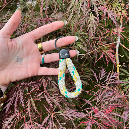 Person hält Sea Shepherd X BRACENET x humanvoll Schlüsselanhänger mit schwarzer Kappe mit dem Sea Shepherd und humanvoll Logo aus weißem Fischereiseil mit gelben und grünen Akzenten vor einen Busch