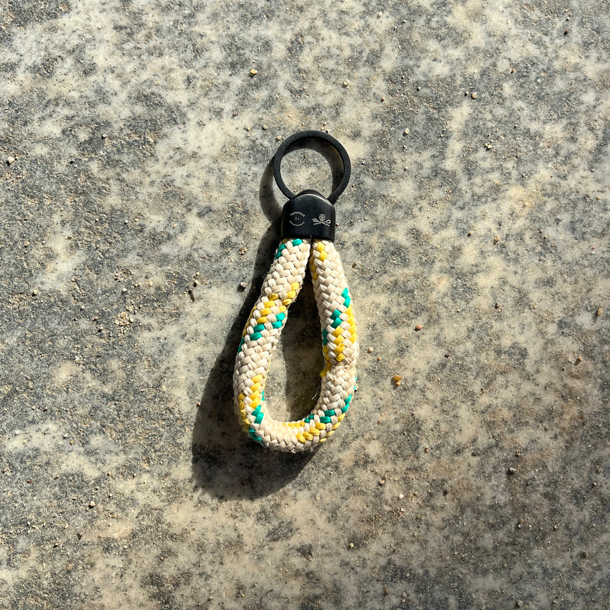 Der Sea Shepherd X BRACENET x humanvoll Schlüsselanhänger mit schwarzer Kappe mit dem Sea Shepherd und humanvoll Logo aus weißem Fischereiseil mit gelben und grünen Akzenten liegt auf grauem Boden