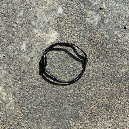 Sea Shepherd X BRACENET x humanvoll Armband aus schwarzen Fischernetzen mit schwarzem Magnetverschluss auf dem das Sea Shepherd Logo, das humanvoll Logo und das BRACENET Logo graviert sind liegt auf grauem Boden