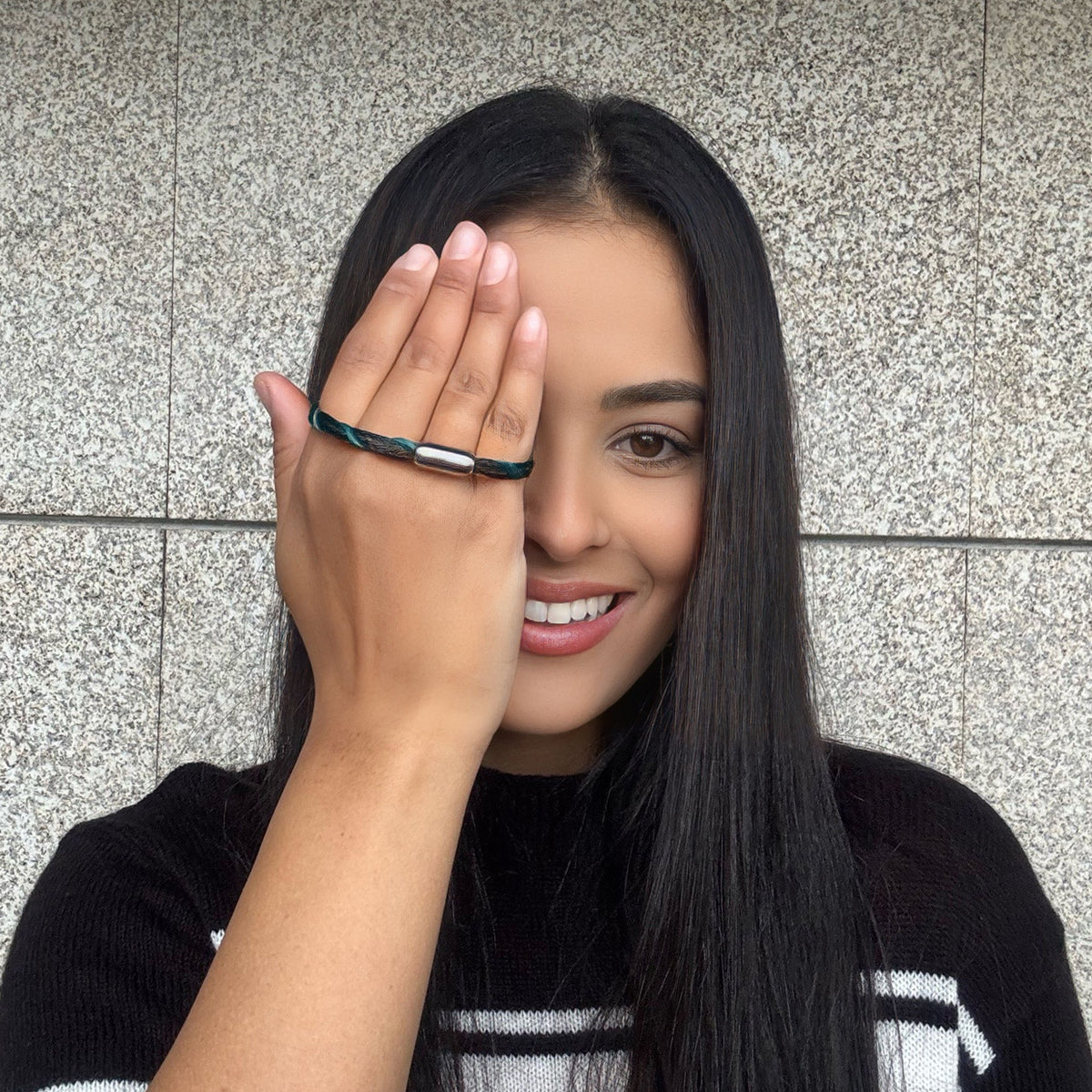 Person hält das November BRACENET Upcycling Armband Timor Sea aus schwarzen und petrolfarbenen Fischernetzen mit silber Verschluss in der Hand und hält sich die Hand vor das Gesicht. 