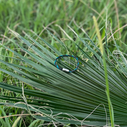 November BRACENET Upcycling Armband Timor Sea aus schwarzen und petrolfarbenen Fischernetzen mit silber Verschluss liegt auf Pflanzen im Grünen.