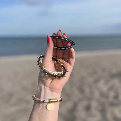 Person hält alle Produkte der BRACENET Glam Edition in der Hand vor dem Meer. Das Armband aus weißem Fischernetz mit goldenen Perlen und goldenem Magnet Verschluss, das Armband aus schwarzem Netz mit goldenen Perlen und goldenem Magnetverschluss, das Armband aus schwarzem Fischernetz mit goldenem Faden durchzogen und goldenem Magnetverschluss und das Armband aus schwarzem Fischernetz mit silbernem Faden durchzogen und silbernem Magnetverschluss.