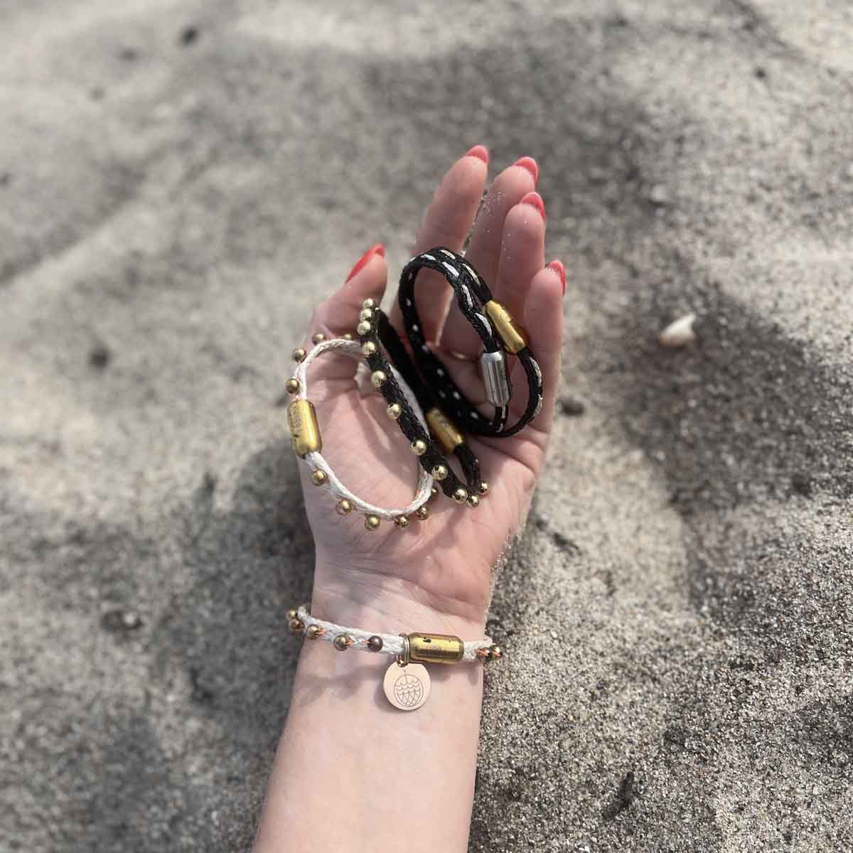 Person hält alle Produkte der BRACENET Glam Edition in der Hand im Sand. Das Armband aus weißem Fischernetz mit goldenen Perlen und goldenem Magnet Verschluss, das Armband aus schwarzem Netz mit goldenen Perlen und goldenem Magnetverschluss, das Armband aus schwarzem Fischernetz mit goldenem Faden durchzogen und goldenem Magnetverschluss und das Armband aus schwarzem Fischernetz mit silbernem Faden durchzogen und silbernem Magnetverschluss.