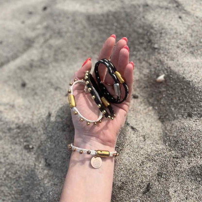 Person hält alle Produkte der BRACENET Glam Edition in der Hand im Sand. Das Armband aus weißem Fischernetz mit goldenen Perlen und goldenem Magnet Verschluss, das Armband aus schwarzem Netz mit goldenen Perlen und goldenem Magnetverschluss, das Armband aus schwarzem Fischernetz mit goldenem Faden durchzogen und goldenem Magnetverschluss und das Armband aus schwarzem Fischernetz mit silbernem Faden durchzogen und silbernem Magnetverschluss.