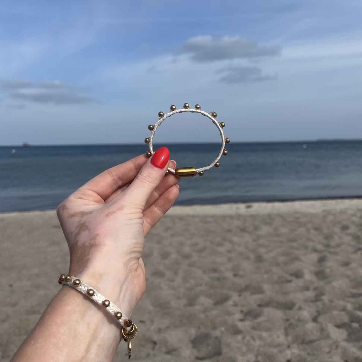 Person hält das BRACENET Glam Edition Armband aus weißem Fischernetz mit goldenen Perlen und goldenem Magnet Verschluss vor das Meer und trägt am Handgelenk ebenfalls die Glam Edition in weiß. 
