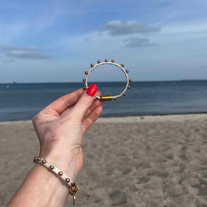 Person hält das BRACENET Glam Edition Armband aus weißem Fischernetz mit goldenen Perlen und goldenem Magnet Verschluss vor das Meer und trägt am Handgelenk ebenfalls die Glam Edition in weiß. 