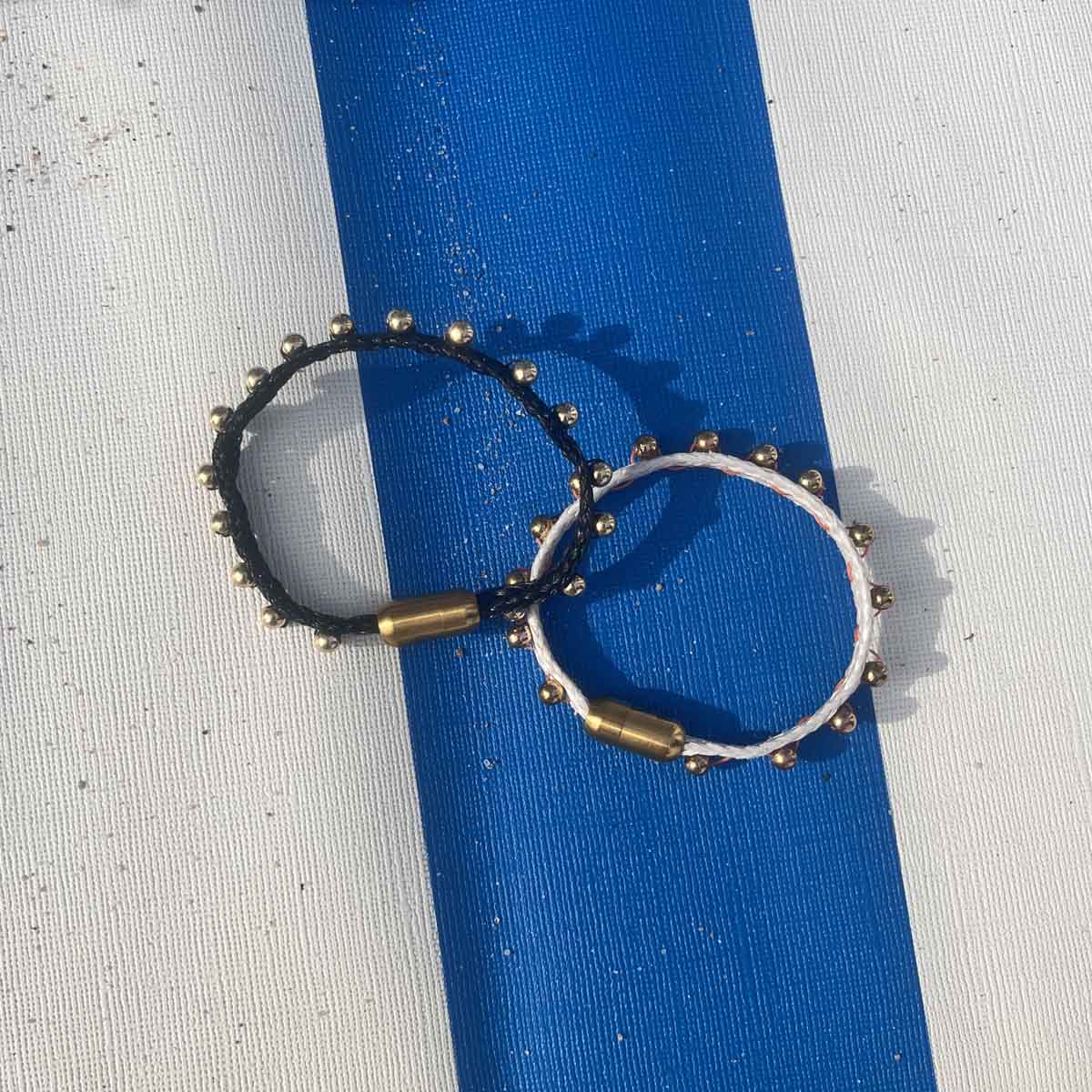 Auf einem blau-weiß gestreiften Strandkorb liegt das BRACENET Glam Edition Armband aus weißem Fischernetz mit goldenen Perlen und goldenem Magnet Verschluss und das BRACENET Glam Edition Armband aus schwarzem Fischernetz mit goldenen Perlen und goldenem Magnet Verschluss.