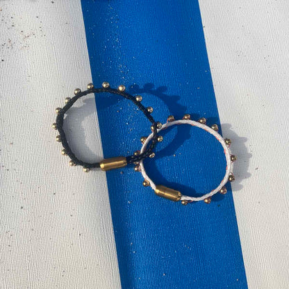 Auf einem blau-weiß gestreiften Strandkorb liegt das BRACENET Glam Edition Armband aus weißem Fischernetz mit goldenen Perlen und goldenem Magnet Verschluss und das BRACENET Glam Edition Armband aus schwarzem Fischernetz mit goldenen Perlen und goldenem Magnet Verschluss.