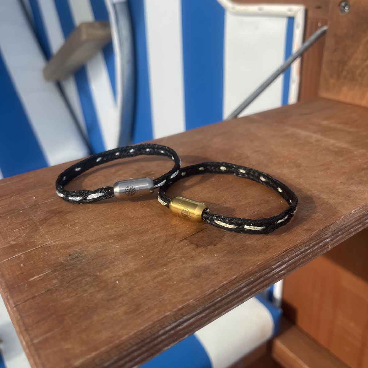 Das BRACENET Glam Edition Armband aus schwarzem Fischernetz mit silbernem Faden durchzogen und silbernem Magnetverschluss und das BRACENET Glam Edition Armband aus schwarzem Fischernetz mit goldenem Faden durchzogen und goldenem Magnetverschluss liegen auf einem Strandkorb-Tisch. 