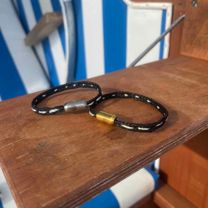 Das BRACENET Glam Edition Armband aus schwarzem Fischernetz mit silbernem Faden durchzogen und silbernem Magnetverschluss und das BRACENET Glam Edition Armband aus schwarzem Fischernetz mit goldenem Faden durchzogen und goldenem Magnetverschluss liegen auf einem Strandkorb-Tisch. 