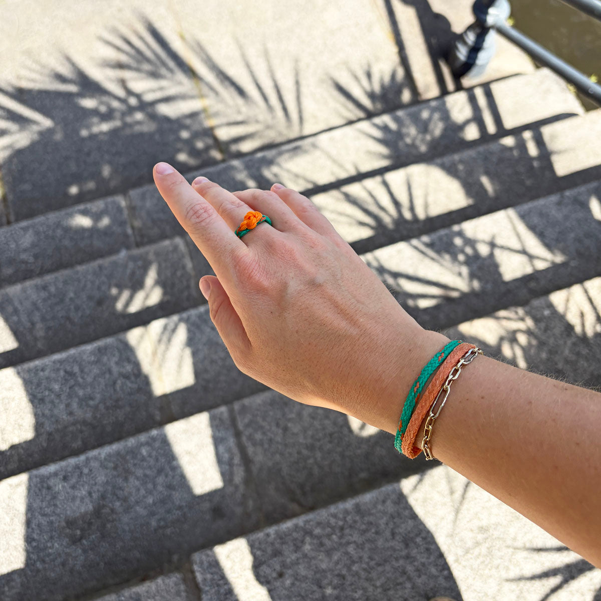 Person trägt August Bracenet Phuket Armband aus Fischernetz mit einem Strang Fischernetz in Grün mit Highlights in Orange, einem Strang Fischernetz in Orange und einem Magnetverschluss in Roségold und Koh Phi Phi Ring aus Fischernetz in Grün und Orange mit Fischernetzknoten in Orange und Endkappen in Silber an der Hand.