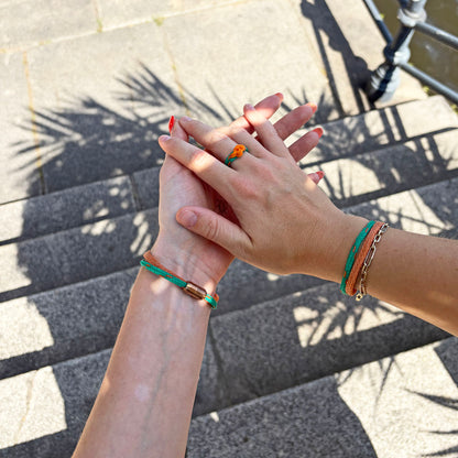 Zwei Personen tragen das August Bracenet Phuket Armband aus Fischernetz mit einem Strang Fischernetz in Grün mit Highlights in Orange, einem Strang Fischernetz in Orange und einem Magnetverschluss in Roségold und August Ring Kohg Phi Phi aus Fischernetz in Grün mit Highlights in Orange und einem orangenen Knoten an der hand und halten die Hände. 