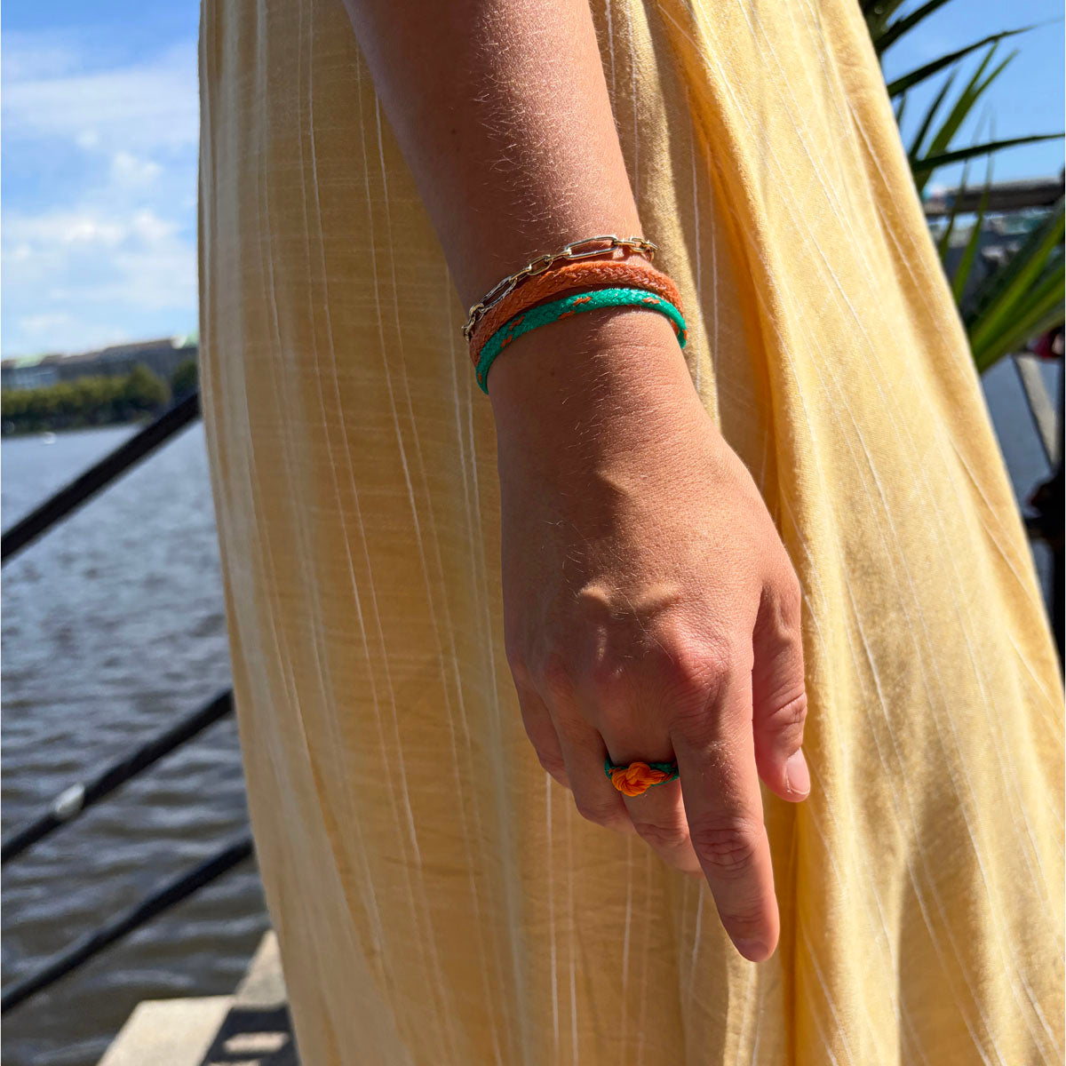 Person trägt August Bracenet Phuket Armband aus Fischernetz mit einem Strang Fischernetz in Grün mit Highlights in Orange, einem Strang Fischernetz in Orange und einem Magnetverschluss in Roségold und August Ring Kohg Phi Phi aus Fischernetz in Grün mit Highlights in Orange und einem orangenen Knoten an der Hand.