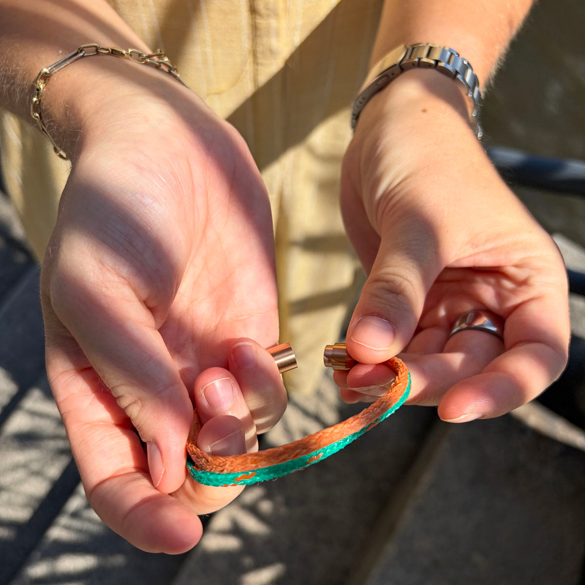 Person öffnet das August Bracenet Phuket Armband aus Fischernetz mit einem Strang Fischernetz in Grün mit Highlights in Orange, einem Strang Fischernetz in Orange und einem Magnetverschluss in Roségold.