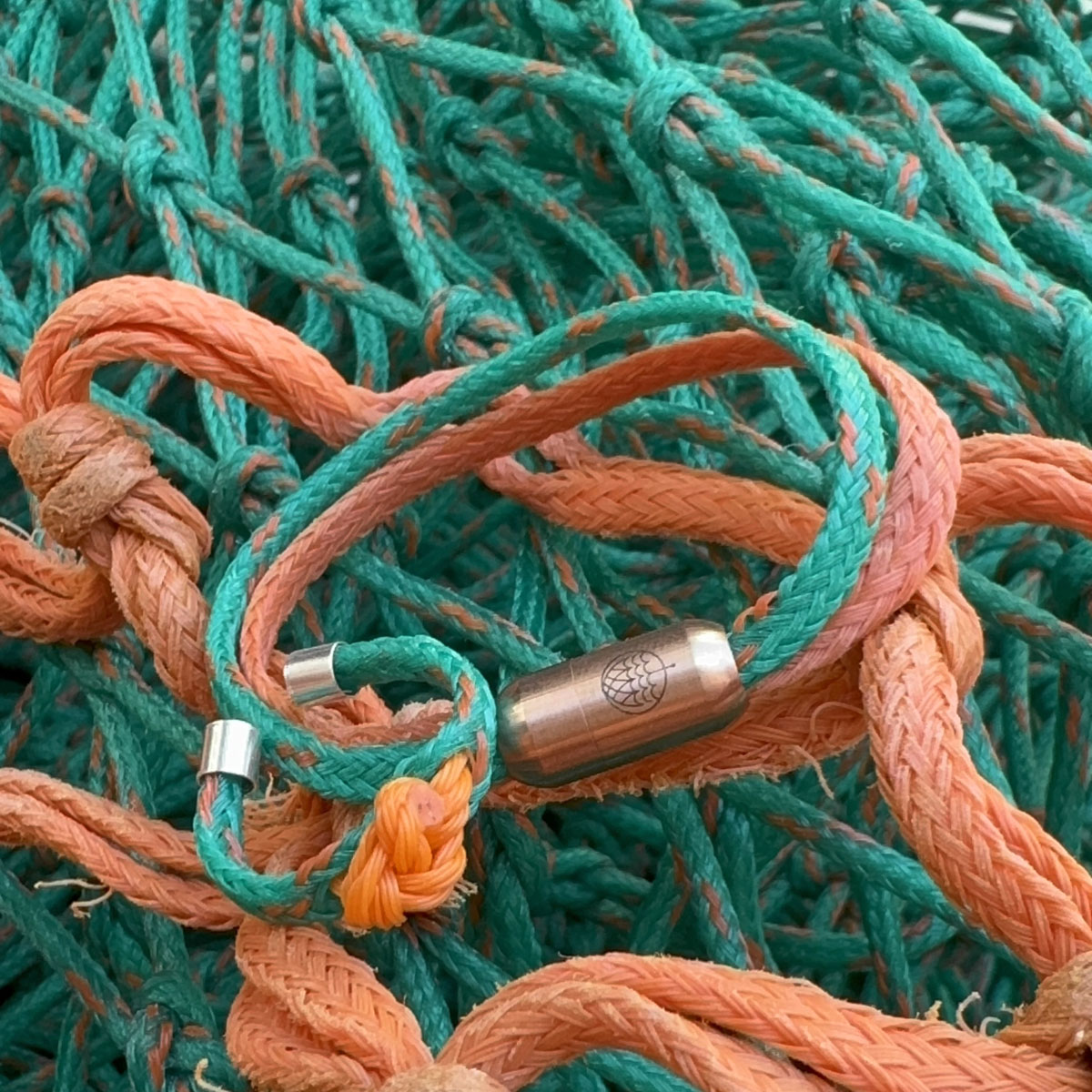 August Bracenet Phuket Armband aus Fischernetz mit einem Strang Fischernetz in Grün mit Highlights in Orange, einem Strang Fischernetz in Orange und einem Magnetverschluss in Roségold und Koh Phi Phi Ring aus Fischernetz in Grün und Orange mit Fischernetzknoten in Orange und Endkappen in Silber, liegt auf Fischernetz. 