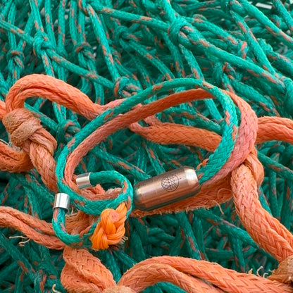 August Bracenet Phuket Armband aus Fischernetz mit einem Strang Fischernetz in Grün mit Highlights in Orange, einem Strang Fischernetz in Orange und einem Magnetverschluss in Roségold und Koh Phi Phi Ring aus Fischernetz in Grün und Orange mit Fischernetzknoten in Orange und Endkappen in Silber, liegt auf Fischernetz. 