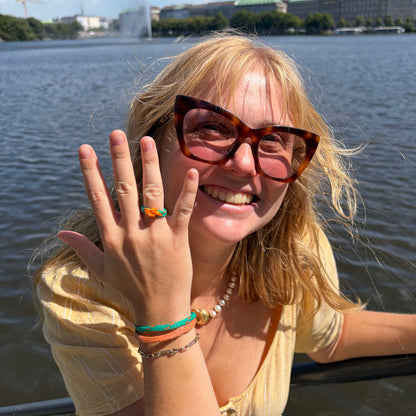 Person trägt August Bracenet Phuket Armband aus Fischernetz mit einem Strang Fischernetz in Grün mit Highlights in Orange, einem Strang Fischernetz in Orange und einem Magnetverschluss in Roségold und August Ring Kohg Phi Phi aus Fischernetz in Grün mit Highlights in Orange und einem orangenen Knoten an der Hand und grinst in die Kamera.