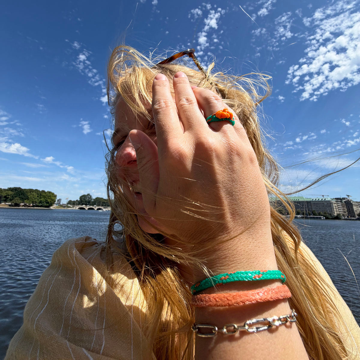 Person trägt August Bracenet Phuket Armband aus Fischernetz mit einem Strang Fischernetz in Grün mit Highlights in Orange, einem Strang Fischernetz in Orange und einem Magnetverschluss in Roségold und August Ring Kohg Phi Phi aus Fischernetz in Grün mit Highlights in Orange und einem orangenen Knoten an der Hand und hält die Hand vor das Gesicht. 