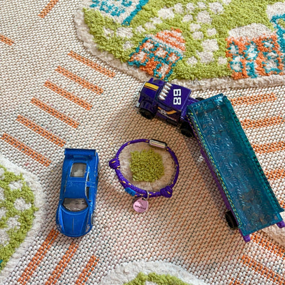 BRACENET Schulkind Bracenet aus Fischernetz in lila mit Makramee-Detil in Blau, Magnetverschluss in Silber und Anhänger in Silber mit Gravur ener Schultüte und der Aufschrift "Schulkind" liegt neben Spielzeugauttos. 