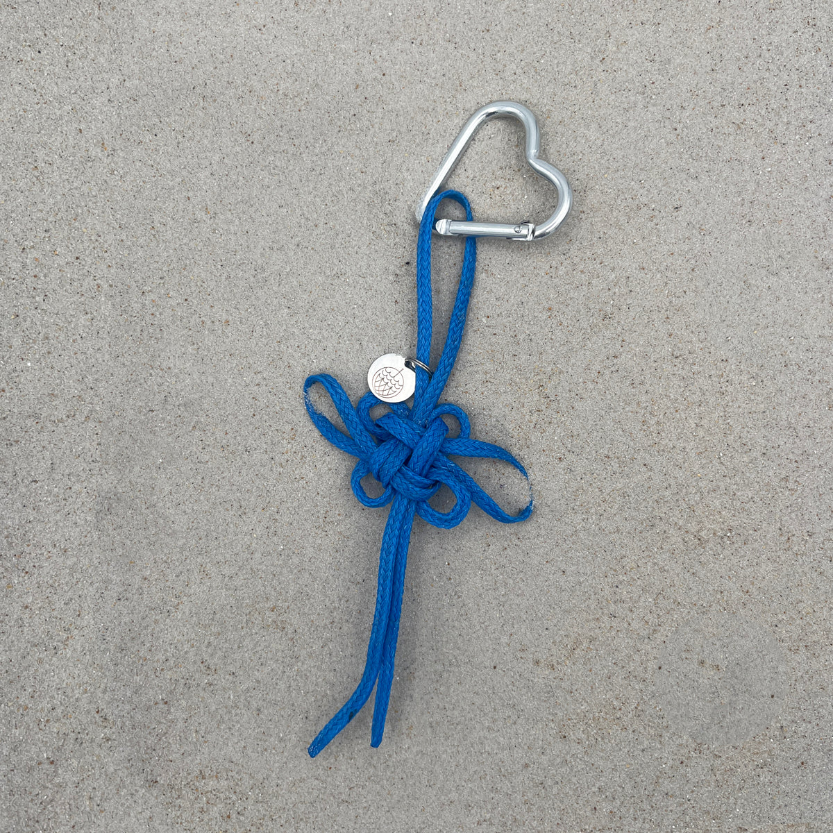 BRACENET Upcycling Taschenanhänger Flores Sea aus blauen alten Fischernetzen mit einem silber Edelstahlanhänger mit BRACENET Logo und einem silber Edelstahlclip in Herzform liegt auf grauem Boden. 