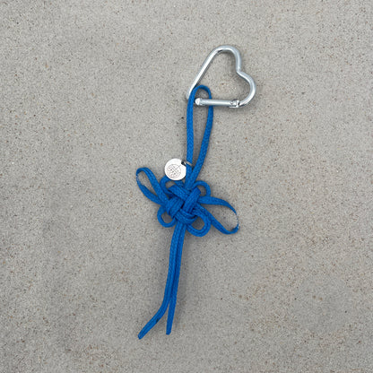 BRACENET Upcycling Taschenanhänger Flores Sea aus blauen alten Fischernetzen mit einem silber Edelstahlanhänger mit BRACENET Logo und einem silber Edelstahlclip in Herzform liegt auf grauem Boden. 