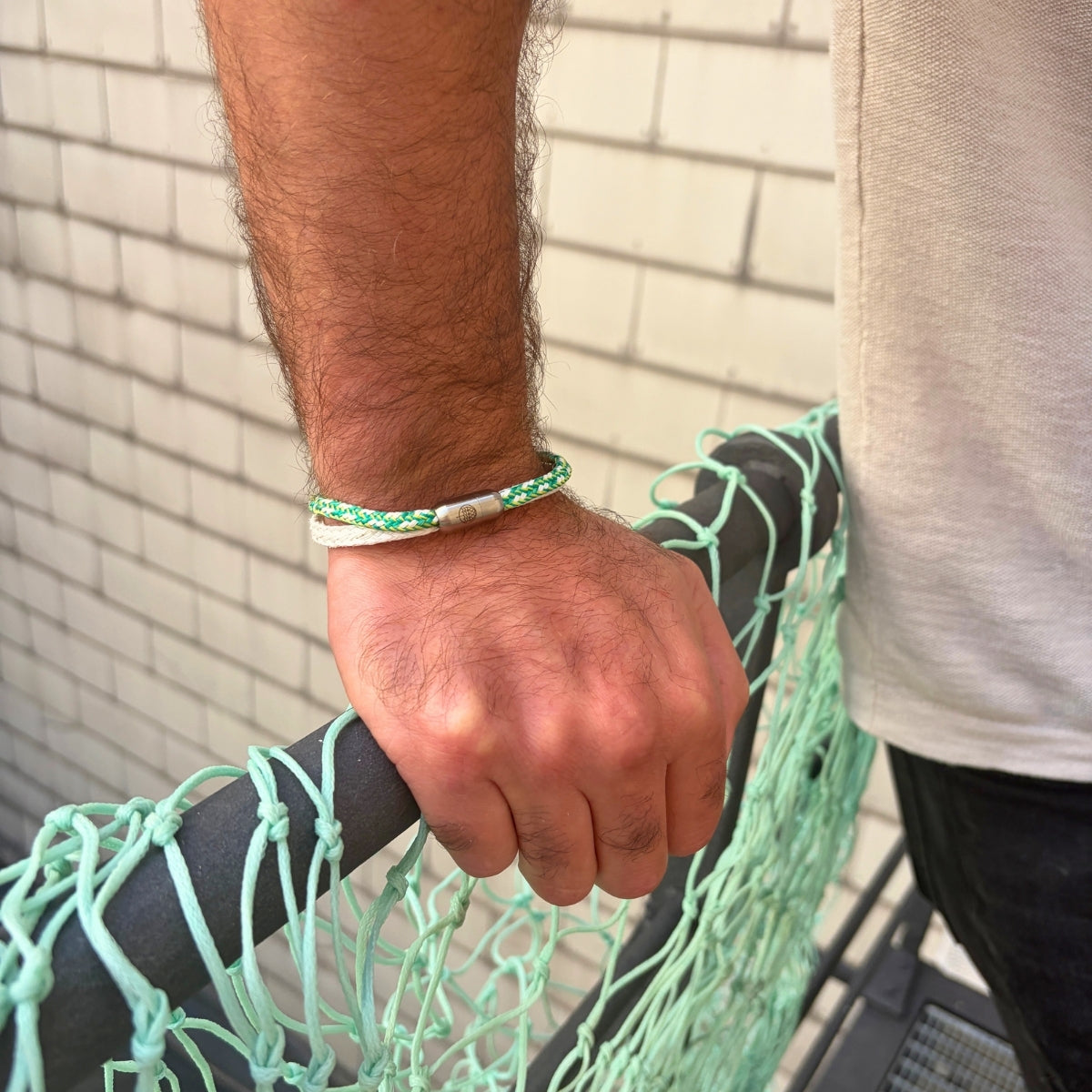 Person trägt das September Bracenet Laguna Verde aus einem Strang Fischernetz in Weiß und einem Strang Fischernetz in Grün, verbunden mit einem Knoten und einem Magnetverschluss in Silber am Handgelenk und stützt sich auf einem Geländer ab.  