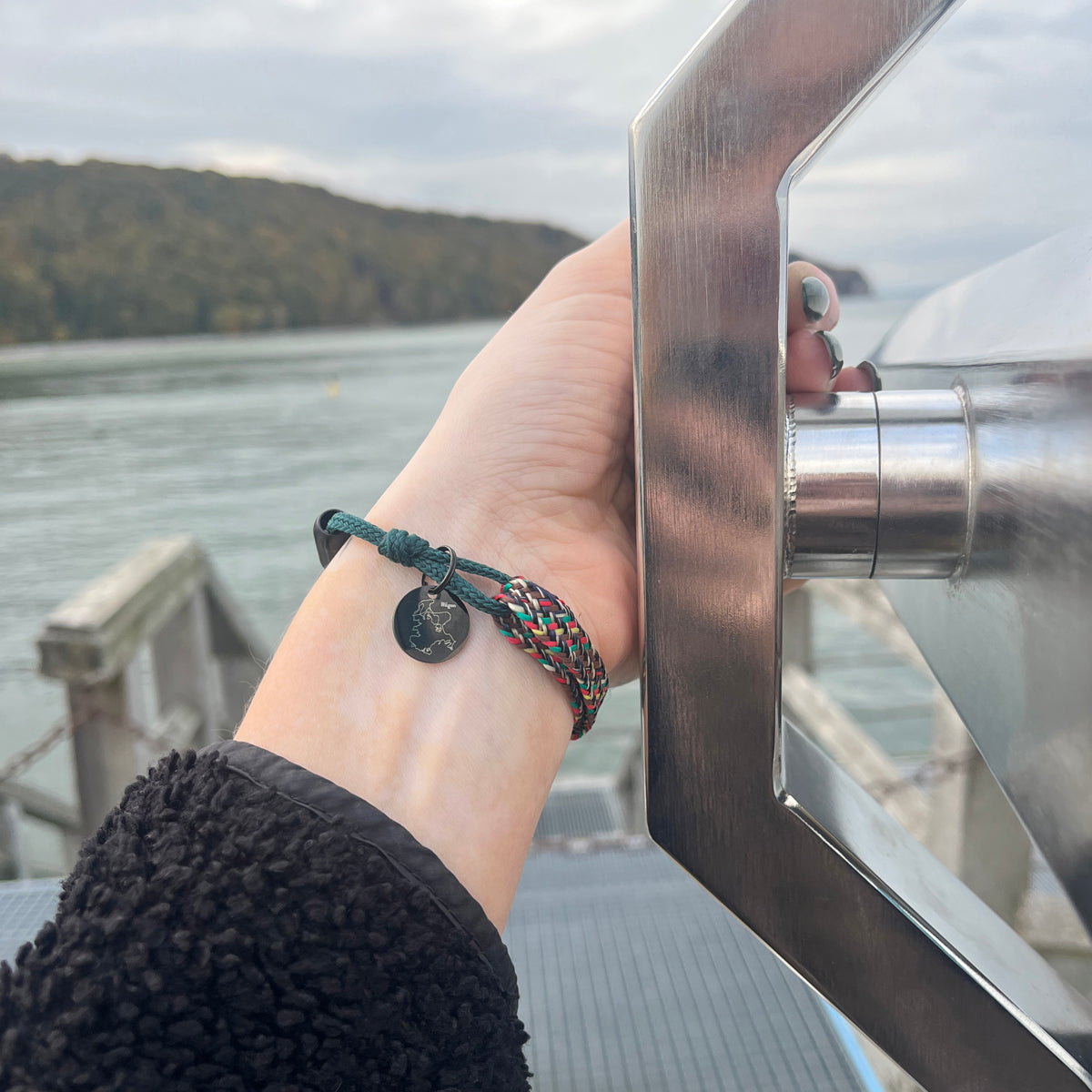 Person trägt das BRACENET Upcycling Armband Insel Rügen aus grünen und bunten Fischernetzen mit schwarzem Verschluss und Anhänger mit Inselgravur am Handgelenk und hält ein Fernrohr fest. 