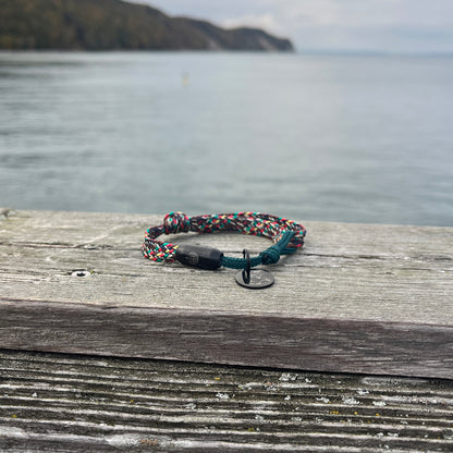BRACENET Upcycling Armband Insel Rügen aus grünen und bunten Fischernetzen mit schwarzem Verschluss und Anhänger mit Inselgravur liegt auf Holz. Im Hintergrund ist das Meer.