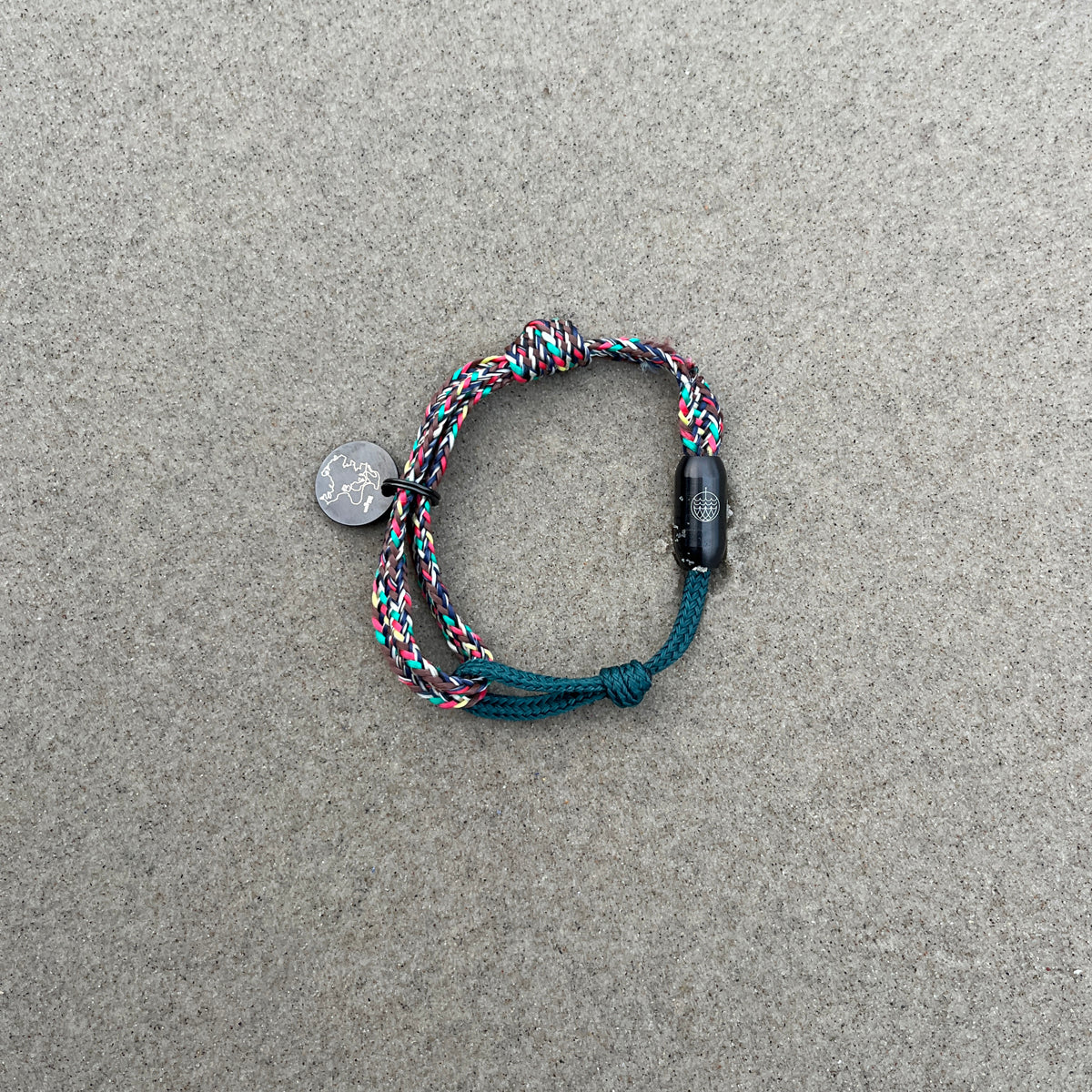 BRACENET Upcycling Armband Insel Rügen aus grünen und bunten Fischernetzen mit schwarzem Verschluss und Anhänger mit Inselgravur liegt im Sand.