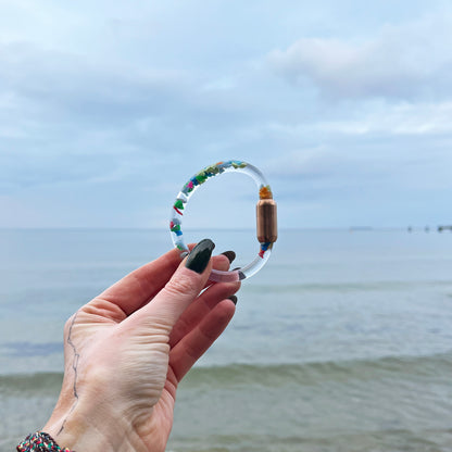 Person hält das BRACENET Floating Upcycling Armband aus alten Plastiktuben gefüllt mit Mikroplastik, destilliertem Wasser und Glycerin mit magnetischem Edelstahlverschluss in rosegold in der Hand. Im Hinterrund sieht man das Meer
