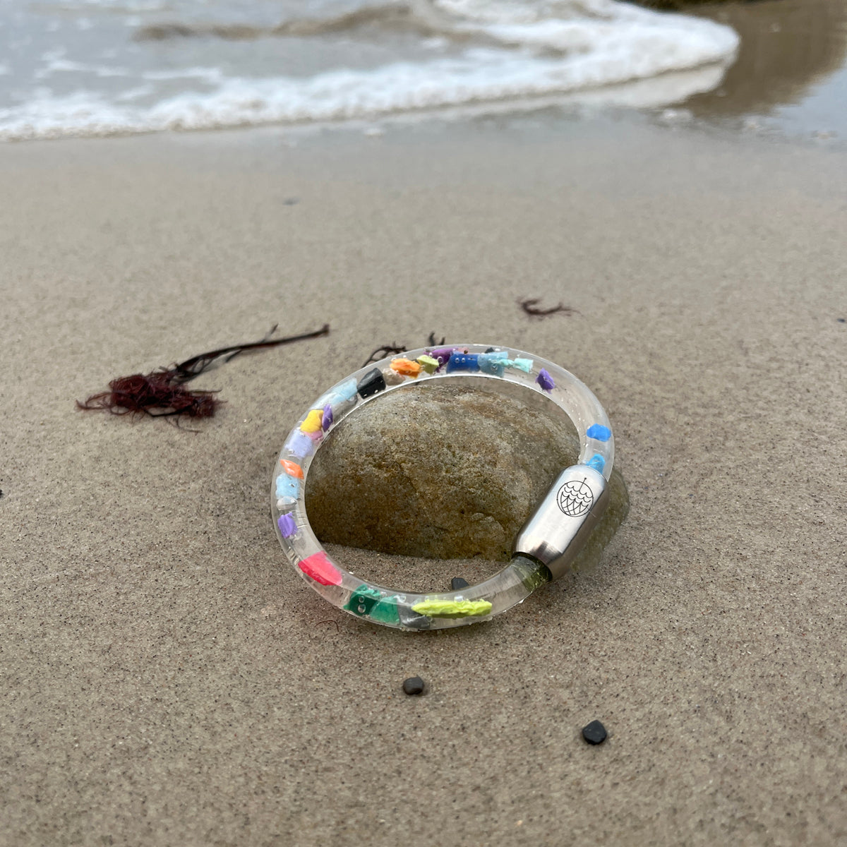 BRACENET Floating Upcycling Armband aus alten Plastiktuben gefüllt mit Mikroplastik, destilliertem Wasser und Glycerin mit magnetischem Edelstahlverschluss in silber liegt auf einem Stein im Sand am Strand