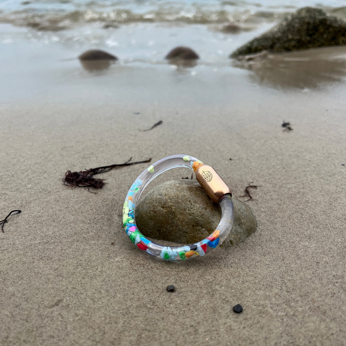 Das BRACENET Floating Upcycling Armband aus alten Plastiktuben gefüllt mit Mikroplastik, destilliertem Wasser und Glycerin mit magnetischem Edelstahlverschluss in rosegold liegt am Strand auf einem Stein im Sand