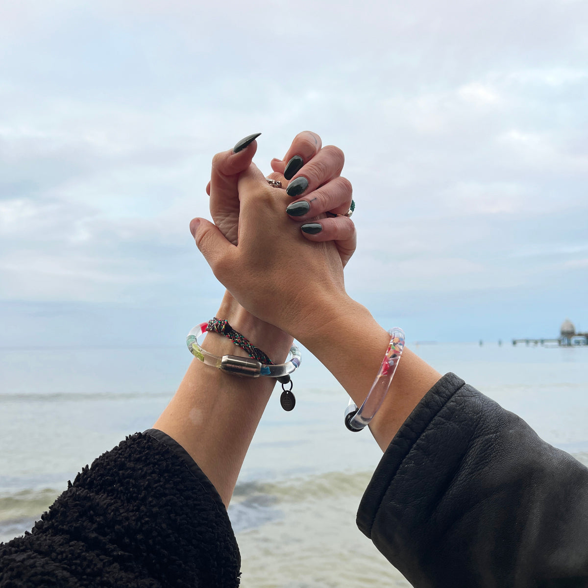 Zwei Personen BRACENET Floating Upcycling Armband aus alten Plastiktuben gefüllt mit Mikroplastik, destilliertem Wasser und Glycerin mit magnetischem Edelstahlverschluss in silber und schwarz und halten sich an den Händen
