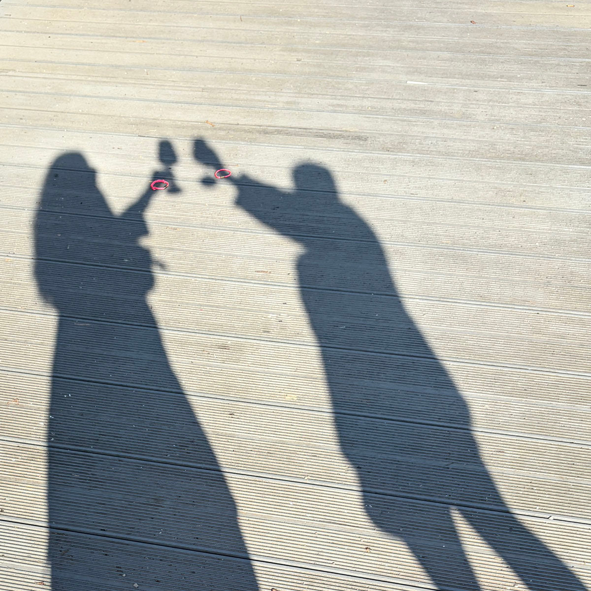 Die Schatten von zwei Personen stoßen an, es liegen zwei Gulf of Bothnia Bracenets aus Fischernetz in Pink mit blauen Punkten an ihren Handgelenken platziert. 
