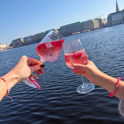 Eine Person trägt das Gulf of Bothnia Bracenet aus Fischernetz in Pink mit blauen Punkten und Magnetverschluss in Roségold am Handgelenk und stößt mit einer anderen Person an, die das Gulf of Bothnia Bracenet aus Fischernetz in Pink mit blauen Punkten und Magnetverschluss Silber am Glas hängen hat. 