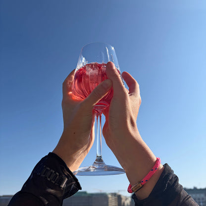 Person trägt das Gulf of Bothnia Bracenet aus Fischernetz in Pink mit blauen Punkten und Magnetverschluss in Roségold am Handgelenk und hält ein Glas in die Luft. 
