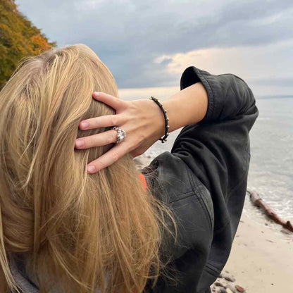 Person trägt das BRACENET Glam Edition Armband aus schwazrm Fischernetz mit goldenen Perlen und goldenem Magnet Verschluss am Handgelenk und fasst sich mit der Hand in die Haare. Die Person steht am Strand. 