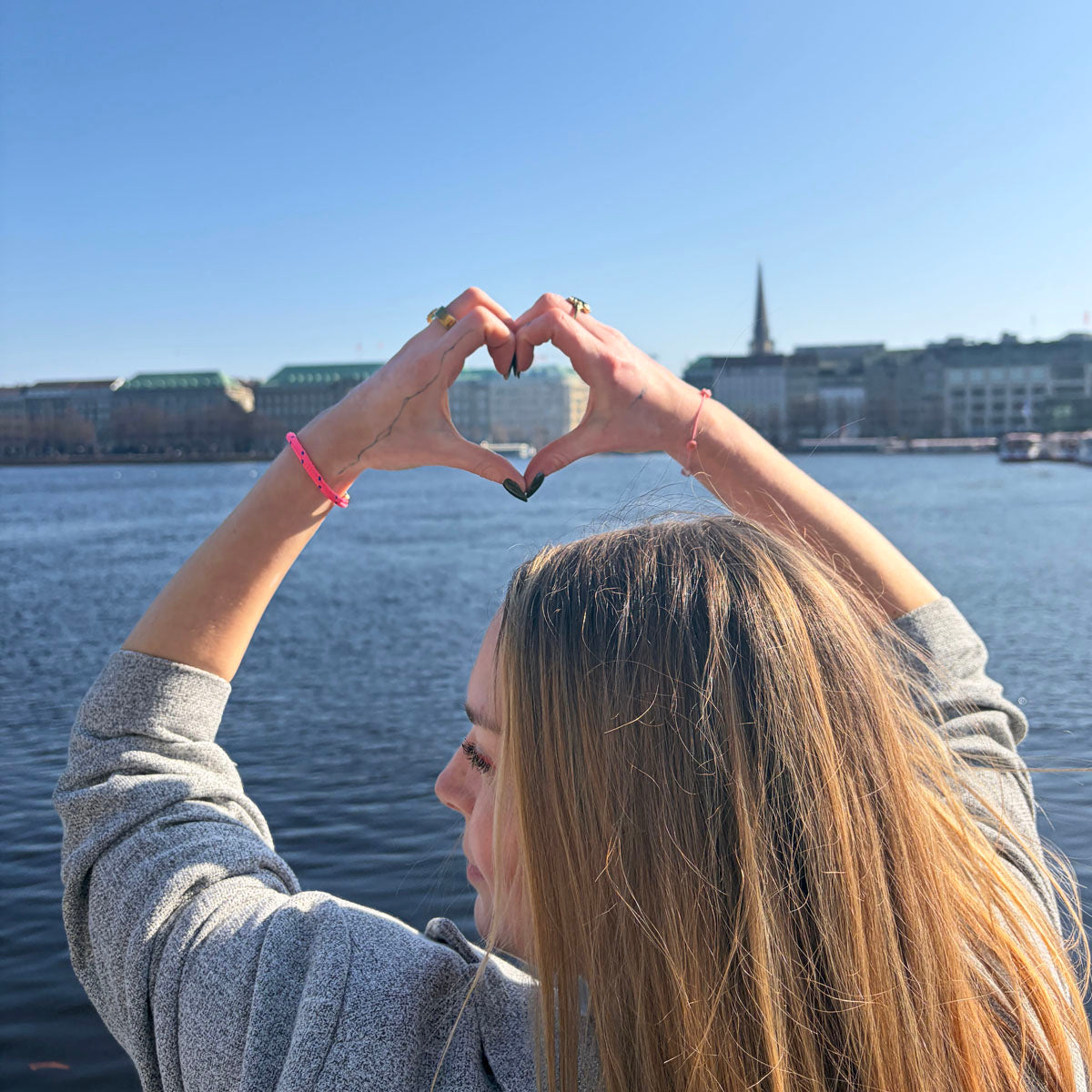 Person trägt das Gulf of Bothnia Bracenet aus Fischernetz in Pink mit blauen Punkten am Handgelenk und zeigt ein Herz in die Luft. 