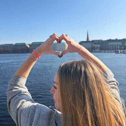 Person trägt das Gulf of Bothnia Bracenet aus Fischernetz in Pink mit blauen Punkten am Handgelenk und zeigt ein Herz in die Luft. 
