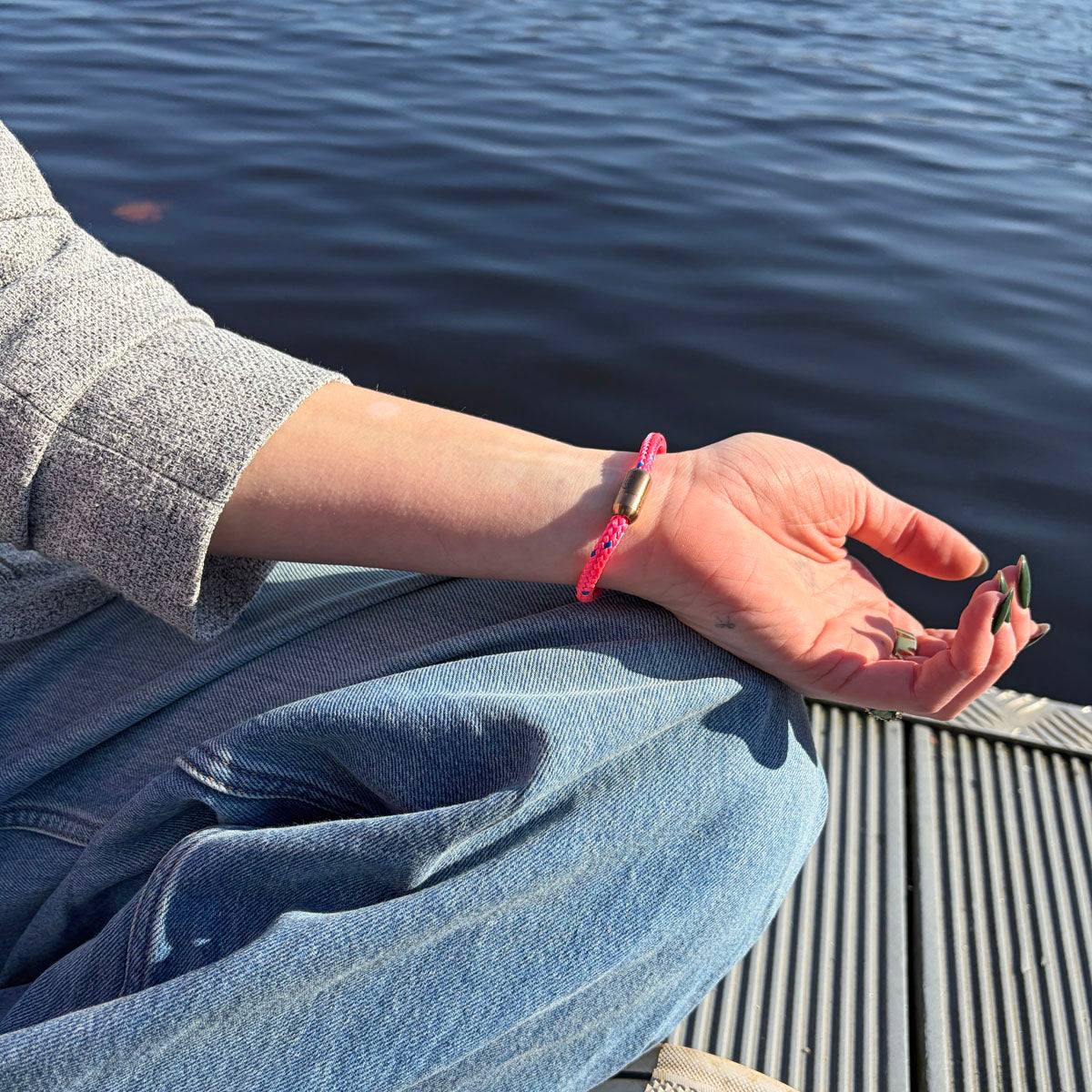 Person trägt das Gulf of Bothnia Bracenet aus Fischernetz in Pink mit blauen Punkten und Magnetverschluss in Roségold am Handgelenk und hat die Hand auf dem Bein liegen. 