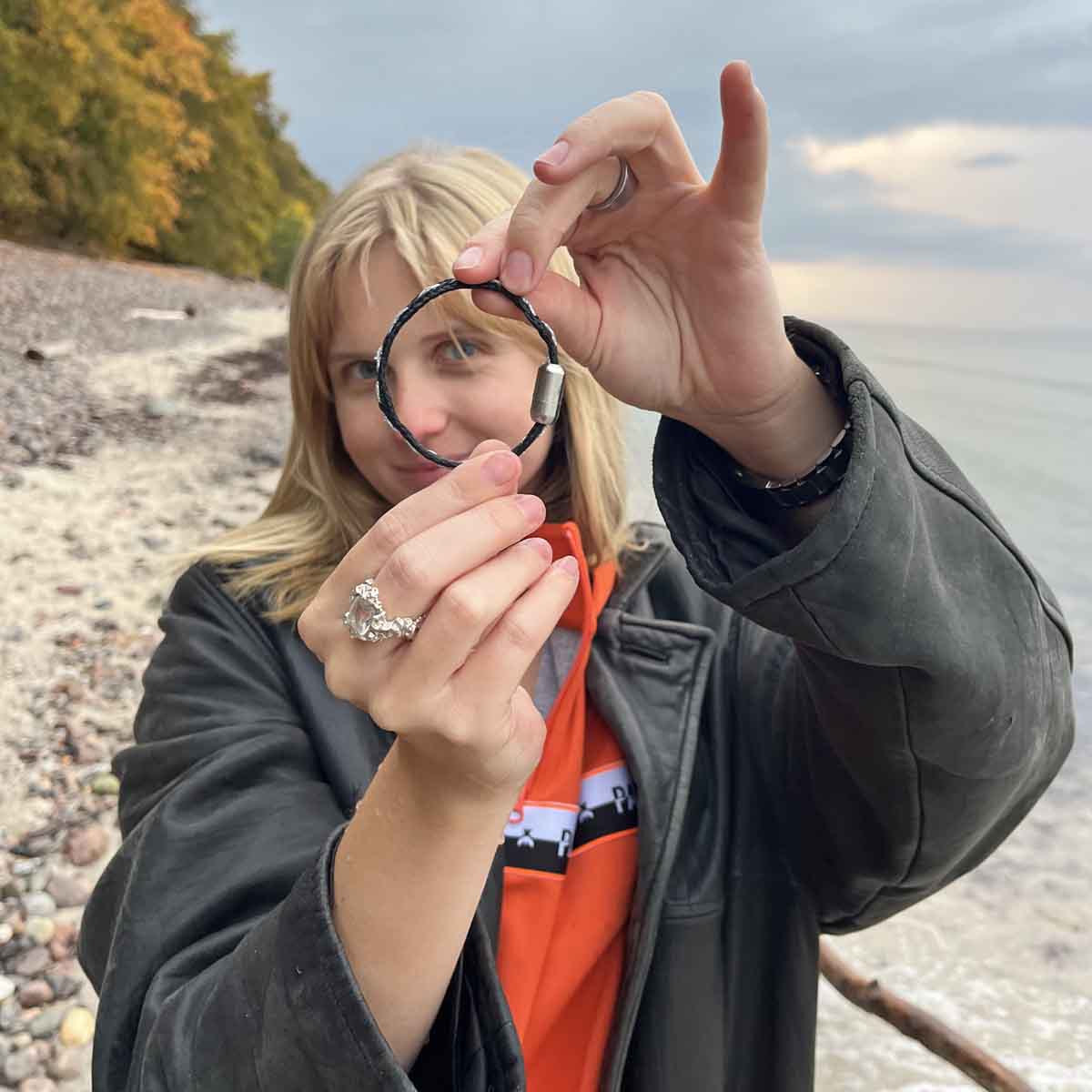 Person hält das BRACENET Glam Edition Armband aus schwarzem Fischernetz mit silbernem Faden durchzogen und silbernem Magnetverschluss vor sich vor ihr Gesicht. Die Person steht am Strand. 