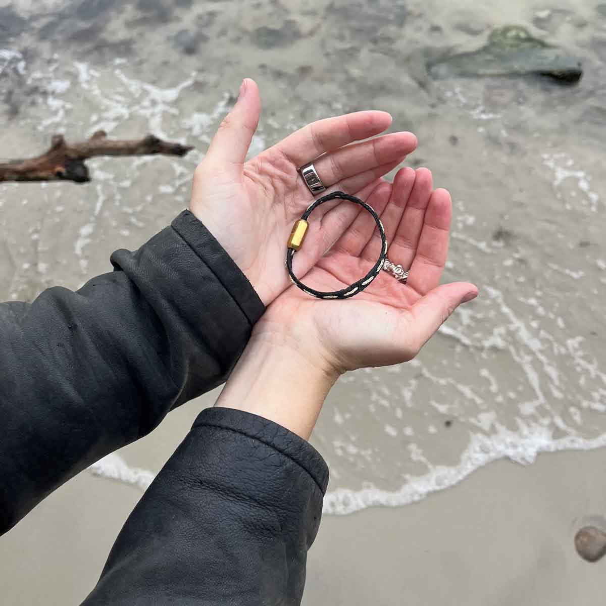 Person hält das BRACENET Glam Edition Armband aus schwarzem Fischernetz mit goldenem Faden durchzogen und goldenem Magnetverschluss in den Händen und hält die Hände über das Meer. 