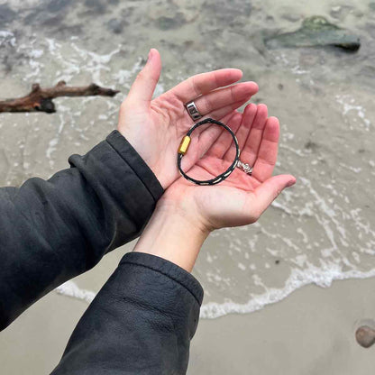 Person hält das BRACENET Glam Edition Armband aus schwarzem Fischernetz mit goldenem Faden durchzogen und goldenem Magnetverschluss in den Händen und hält die Hände über das Meer. 