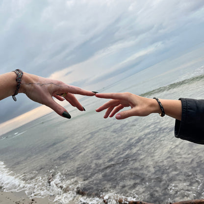 Eine Personen trägt das BRACENET Upcycling Armband Insel Rügen aus grünen und bunten Fischernetzen mit schwarzem Verschluss und Anhänger mit Inselgravur am Handgelenk und berührt mit dem Finger die Hand einer anderen Person. 