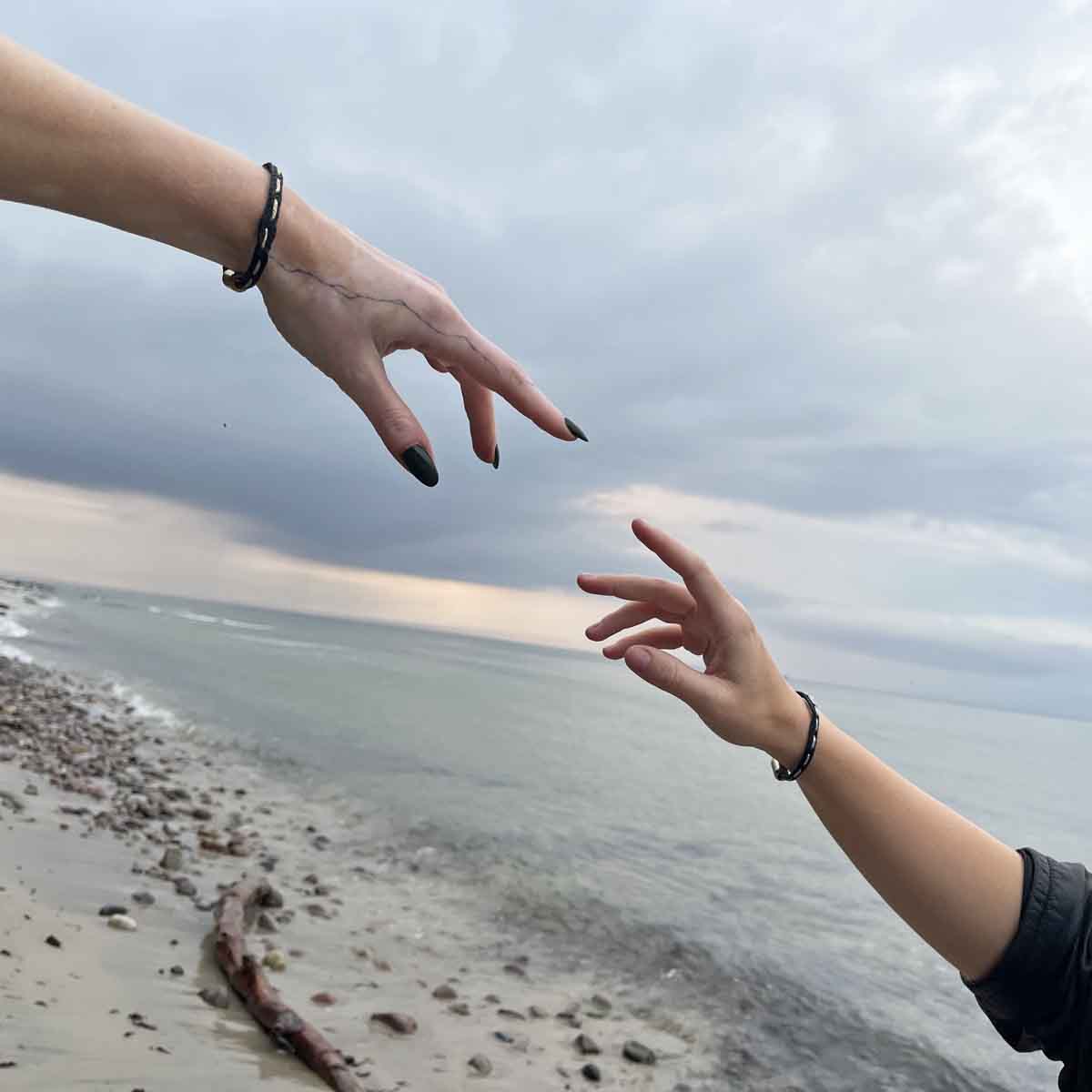 Zwei Personen strecken die Finger aufeinander zu. An den Handgelenk trägt eine Person das BRACENET Glam Edition Armband aus schwarzem Fischernetz mit goldenem Faden durchzogen und goldenem Magnetverschluss und die andere das BRACENET Glam Edition Armband aus schwarzem Fischernetz mit silbernem Faden durchzogen und silbernem Magnetverschluss.