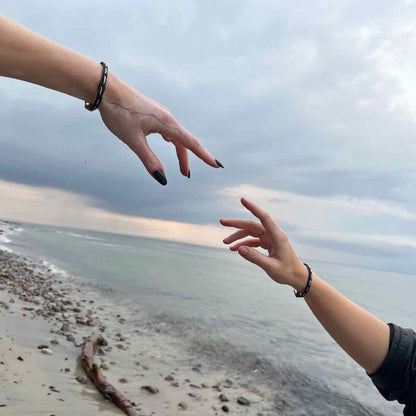 Zwei Personen strecken die Finger aufeinander zu. An den Handgelenk trägt eine Person das BRACENET Glam Edition Armband aus schwarzem Fischernetz mit goldenem Faden durchzogen und goldenem Magnetverschluss und die andere das BRACENET Glam Edition Armband aus schwarzem Fischernetz mit silbernem Faden durchzogen und silbernem Magnetverschluss.